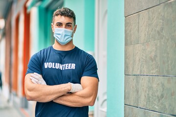 Young hispanic volunteer man with arms crossed wearing medical mask at the city.