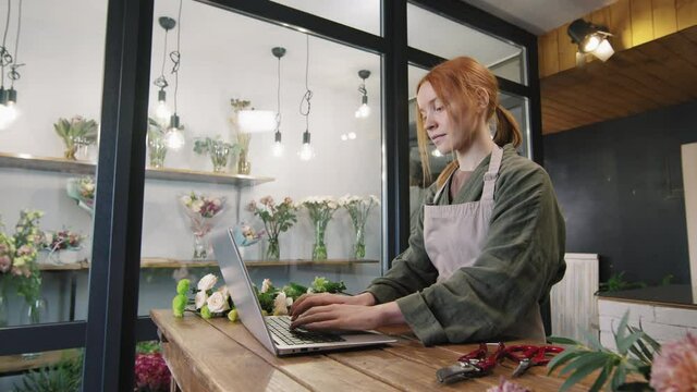 Panning medium shot of young woman typing on laptop at counter of small flower shop, while her colleague placing flowers at store window
