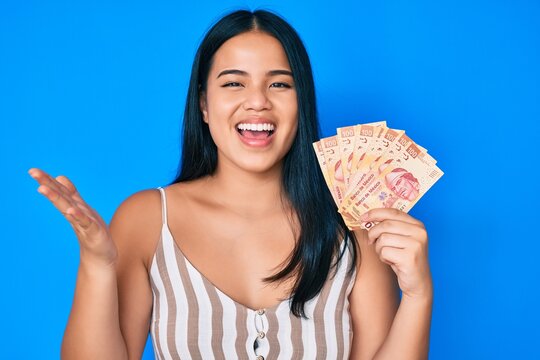 Young beautiful asian girl holding mexican pesos celebrating achievement with happy smile and winner expression with raised hand