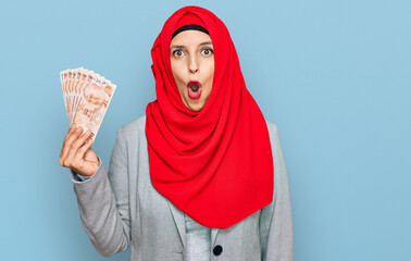 Beautiful hispanic woman wearing islamic hijab holding turkish lira banknotes scared and amazed with open mouth for surprise, disbelief face