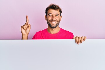 Handsome man with beard holding blank empty banner smiling with an idea or question pointing finger with happy face, number one