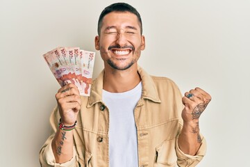 Handsome man with tattoos holding 10 colombian pesos banknotes screaming proud, celebrating victory and success very excited with raised arm - obrazy, fototapety, plakaty