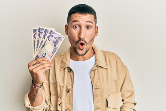 Handsome man with tattoos holding japanese yen banknotes scared and amazed with open mouth for surprise, disbelief face