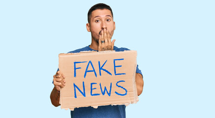 Handsome man with tattoos holding fake news banner covering mouth with hand, shocked and afraid for...