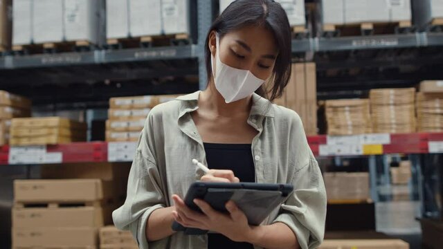 Young Asia businesswoman manager wear face mask looking for goods using digital tablet checking inventory levels stand in retail shopping center. Distribution, Logistics, Packages ready for shipment.