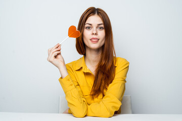 cheerful red-haired woman sits at the table in a yellow shirt with a heart-shaped lollipop