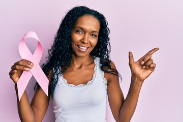 Middle age african american woman holding pink cancer ribbon smiling happy pointing with hand and...