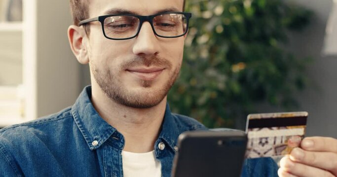 Close up portrait of happy caucasian male shopper with bank card and mobile phone sitting at home on sofa.
