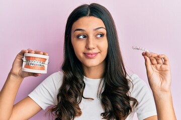 Young hispanic woman holding invisible aligner orthodontic and braces smiling looking to the side and staring away thinking.