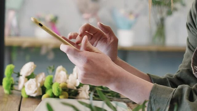 Tilting-up close-up of unrecognizable female hands of florist scrolling feed on smartphone while standing at counter in small flower shop