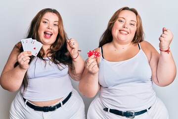 Plus size caucasian sisters woman holding poker chips and cards screaming proud, celebrating victory and success very excited with raised arms