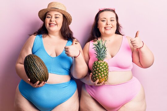 Young plus size twins wearing bikini holding melon and pineapple smiling happy and positive, thumb up doing excellent and approval sign