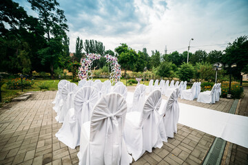 decoration of an outdoor ceremony in a green garden, an arch of fresh flowers, a white path for newlyweds, chairs in white covers