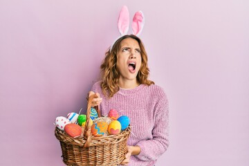 Young caucasian woman wearing cute easter bunny ears holding colored egg angry and mad screaming frustrated and furious, shouting with anger looking up.