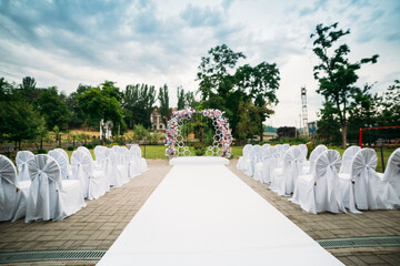 decoration of an outdoor ceremony in a green garden, an arch of fresh flowers, a white path for newlyweds, chairs in white covers