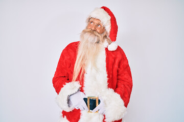 Old senior man with grey hair and long beard wearing santa claus costume with suspenders smiling looking to the side and staring away thinking.