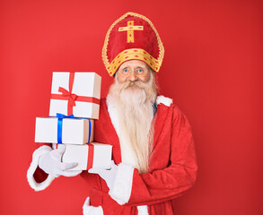 Old senior man with grey hair and long beard wearing saint nicholas costume holding presents looking positive and happy standing and smiling with a confident smile showing teeth