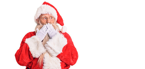 Old senior man with grey hair and long beard wearing traditional santa claus costume laughing and embarrassed giggle covering mouth with hands, gossip and scandal concept