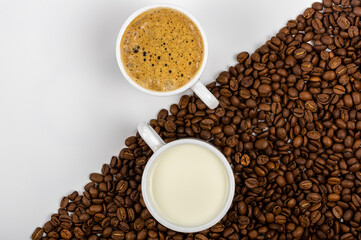 A cup of coffee with froth and a cup of milk standing on coffee beans scattered on a white background.
