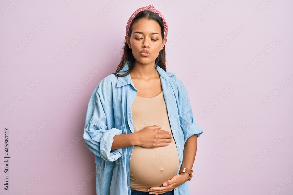 Sticker Beautiful hispanic woman expecting a baby, touching pregnant belly making fish face with mouth and squinting eyes, crazy and comical.