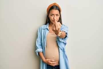 Beautiful hispanic woman expecting a baby, touching pregnant belly showing middle finger, impolite...