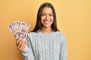 Beautiful hispanic woman holding 500 mexican pesos banknotes looking positive and happy standing and smiling with a confident smile showing teeth