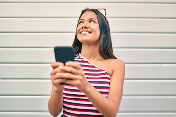 Young latin girl smiling happy using smartphone at the city.