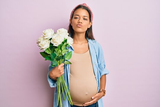 Beautiful hispanic woman expecting a baby holding flowers looking at the camera blowing a kiss being lovely and sexy. love expression.