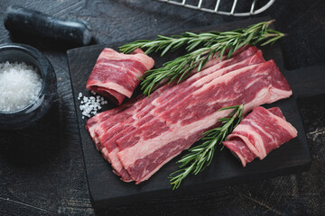 Raw marbled beef bacon with fresh rosemary and salt on a black wooden chopping board, horizontal shot