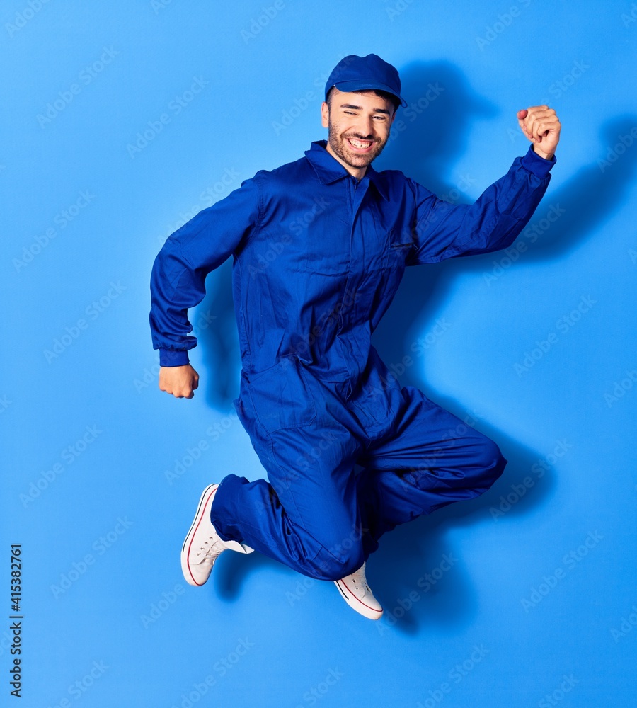 Wall mural Young handsome hispanic man wearing painter uniform and cap smiling happy. Jumping with smile on face doig winner sign with fists up over isolated blue background