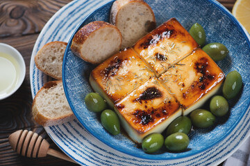 Blue bowl with baked feta cheese, green olives and baguette, studio shot on a brown rustic wooden...