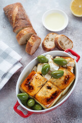 Baked feta cheese served with green olives, bread and olive oil, vertical shot on a beige stone background