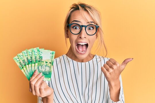 Beautiful blonde woman holding south african 10 rand banknotes pointing thumb up to the side smiling happy with open mouth
