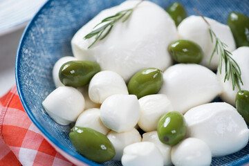 Mozzarella balls of different sizes, green olives and fresh rosemary in a blue bowl, close-up, selective focus