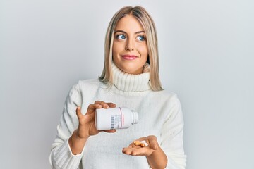 Beautiful blonde woman holding pills smiling looking to the side and staring away thinking.