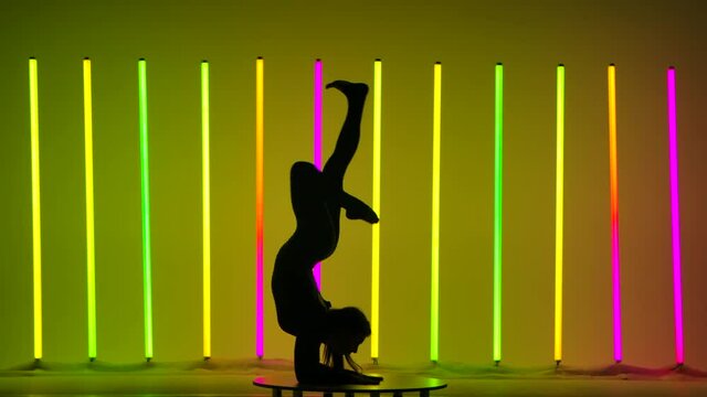 The gymnast moves her legs during handstand and cheststand, then sits on the split and demonstrates stretching. The girl in the studio against the background of multicolored neon lamps. Close up.