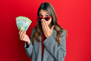 Young brunette woman holding russian 200 ruble banknotes covering mouth with hand, shocked and afraid for mistake. surprised expression