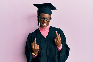 Young african american girl wearing graduation cap and ceremony robe showing middle finger doing...