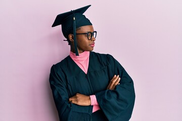 Young african american girl wearing graduation cap and ceremony robe looking to the side with arms crossed convinced and confident