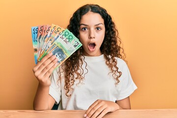 Teenager hispanic girl holding australian dollars scared and amazed with open mouth for surprise, disbelief face