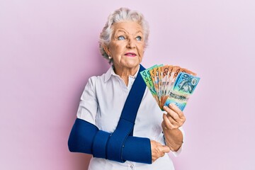 Senior grey-haired woman wearing arm on sling holding australia dollars banknotes smiling looking...