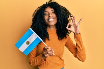 African american woman with afro hair holding argentina flag doing ok sign with fingers, smiling friendly gesturing excellent symbol