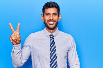 Young latin man wearing business clothes smiling with happy face winking at the camera doing victory sign. number two.
