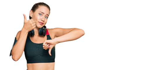 Beautiful blonde woman wearing gym clothes and using headphones doing thumbs up and down,...