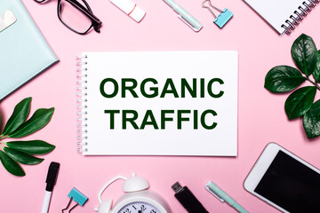 Fototapeta na wymiar ORGANIC TRAFFIC is written in a white notebook on a pink background surrounded by business accessories and green leaves.