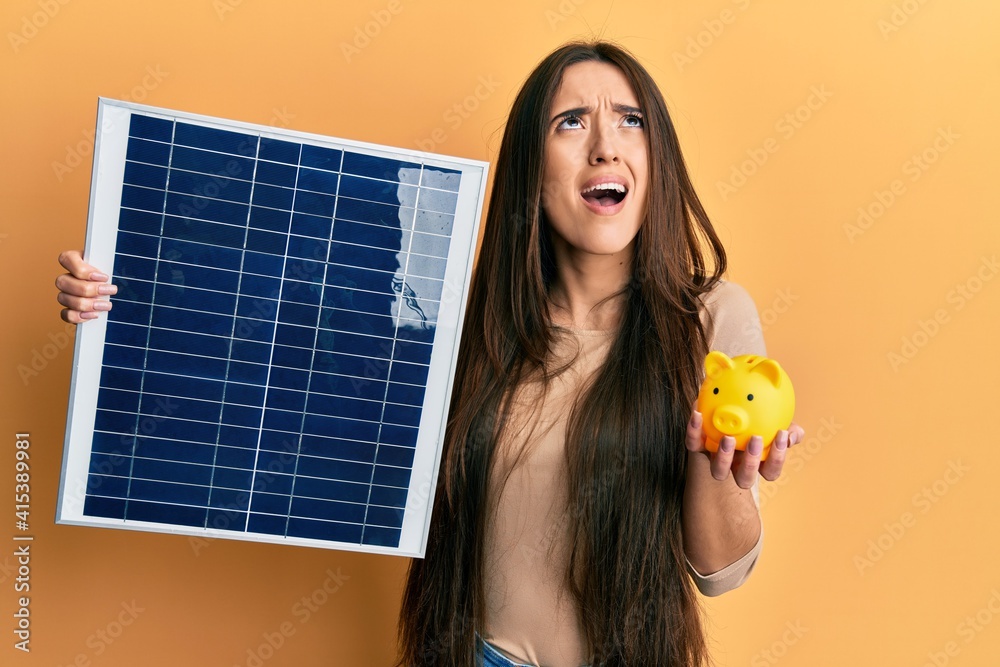 Poster young hispanic girl holding photovoltaic solar panel and piggy bank angry and mad screaming frustrat