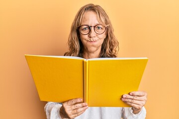 Middle age blonde woman reading a book wearing glasses puffing cheeks with funny face. mouth inflated with air, catching air.