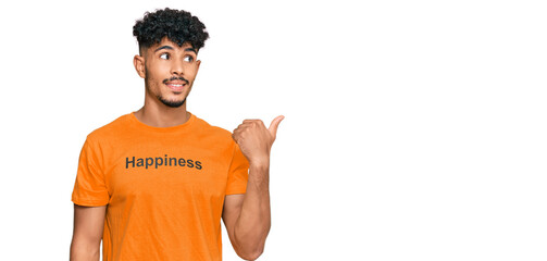 Young arab man wearing tshirt with happiness word message smiling with happy face looking and pointing to the side with thumb up.