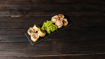 Custom sushi roll in tempura with nori, fresh salmon, tuna, avocado, masago caviar, drizzled with pineapple sauce with salad pouring as decoration on a black plate on a wooden table and background.