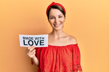 Young brunette woman with short hair holding made with love banner looking positive and happy standing and smiling with a confident smile showing teeth